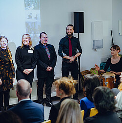 Chormitglieder stehen nebeneinander vor dem Publikum, eine Percussionistin sitzt daneben und spielt. Die Zuhörenden verfolgen die Darbietung aufmerksam.