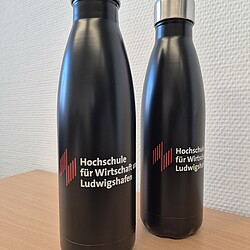Zwei schwarze Edelstahl-Thermosflaschen mit silbernem Schraubverschluss stehen nebeneinander. Beide Flaschen tragen das Hochschullogo in Weiß und Rot.