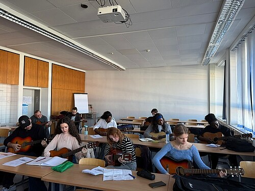 Mehrere Teilnehmende sitzen in einem Klassenraum an Tischen und spielen gemeinsam Gitarre. Vor ihnen liegen Notenblätter, während sie konzentriert üben und die Sonne durch große Fenster scheint.