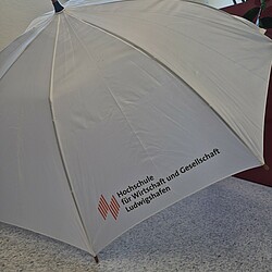 Ein weißer Stockschirm mit Holzgriff und dem Logo der Hochschule für Wirtschaft und Gesellschaft Ludwigshafen ist geöffnet auf dem Boden aufgestellt. Im Hintergrund ist ein Fenster mit blauem Himmel zu sehen.