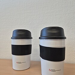 Zwei weiße Thermobecher mit schwarzem Deckel und gummiertem Griff stehen nebeneinander auf einem Tisch. Beide Becher tragen das Logo der Hochschule in kleiner roter Schrift.