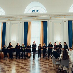 Der Hochschulchor steht in einer Reihe vor einem sitzenden Publikum in einem festlichen Raum mit hohen Fenstern und blauen sowie goldenen Vorhängen und singt gemeinsam.