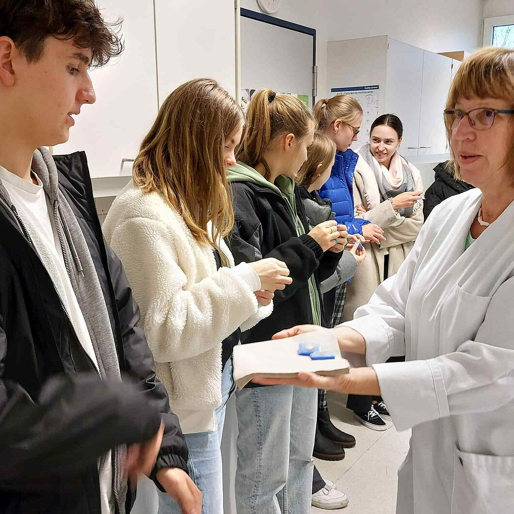 Eine Gruppe von Schüler*innen erhält von einer Frau im Laborkittel eine praktische Einführung in ein medizinisches oder naturwissenschaftliches Thema.