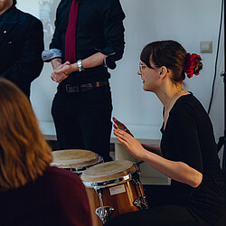 Eine junge Frau sitzt seitlich an einer Wand und spielt konzentriert auf zwei Congas. Im Vordergrund sind Zuhörende zu sehen.