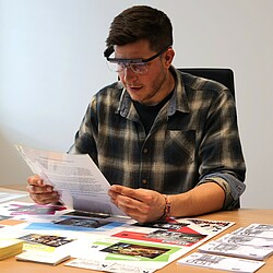 Ein Mann trägt eine Eyetracking-Brille und liest ein Dokument, während er an einem Planspiel teilnimmt, das den Besuch eines Einkaufszentrums simuliert. Vor ihm auf dem Tisch sind verschiedene Geschäfte und Bereiche des Einkaufszentrums auf einer Spielfläche abgebildet