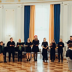 Der Hochschulchor singt in einer Reihe vor einem sitzenden Publikum in einem festlichen Saal. Links sind Conga-Trommeln und eine Cajón zu sehen, die von einem Chormitglied gespielt werden. Die Sängerinnen und Sänger tragen schwarze Kleidung, während eine Sängerin ein rotes Barett trägt.