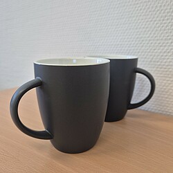 Zwei matt-schwarze Kaffeetassen mit Henkel stehen nebeneinander auf einem Tisch. Die Innenseite der Tassen ist weiß, ein Logo ist außen nicht sichtbar.