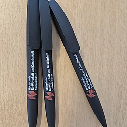Drei schwarze Kugelschreiber mit Softtouch-Oberfläche und dem Logo der Hochschule für Wirtschaft und Gesellschaft Ludwigshafen liegen fächerförmig auf einem Tisch. Die Stifte haben eine schlichte, moderne Optik.
