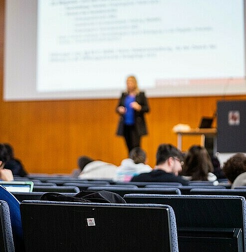 Blick von hinten über Köpfe in Stuhlreihen hinweg auf eine Lehrperson vor großer Leinwand