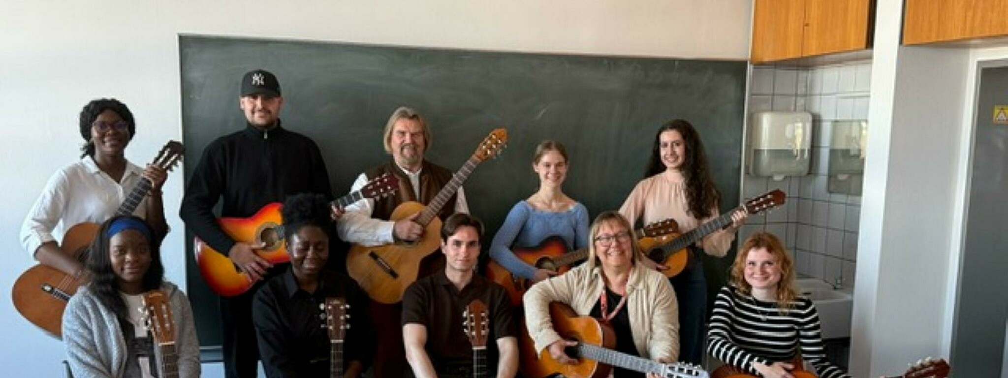 Elf Personen mit Gitarren posieren gemeinsam in einem Klassenraum für ein Gruppenfoto. Sie sitzen und stehen vor einer Tafel und lächeln in die Kamera.
