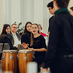 Der Chor singt in einem festlichen Saal, während eine Frau in der Nähe die Trommeln im Tagt schlägt.