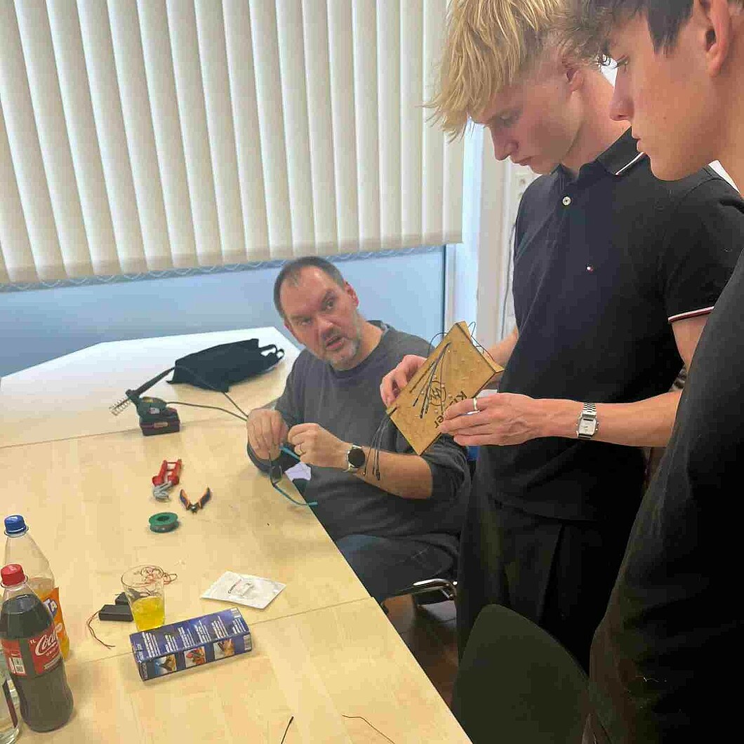 Eine Gruppe junger Menschen baut mit technischer Unterstützung ein elektronisches Projekt mit Kabeln und Lötmaterial zusammen.