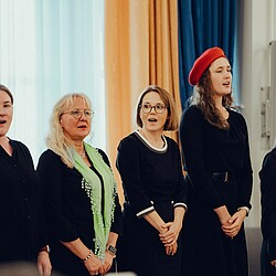 Fünf Chorsängerinnen, die zusammen singen, eine von ihnen trägt ein rotes Barett und eine andere ein grünes Tuch.