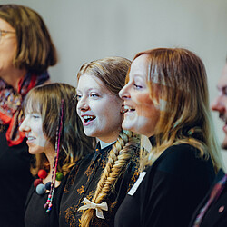Mehrere Chormitglieder singen mit Freude, einige lachen während des Auftritts. Die Sängerinnen und Sänger sind festlich gekleidet und stehen nah beieinander.