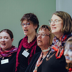 Vier Sängerinnen singen mit offenem Mund, tragen farbige Tücher und Namensschilder. Sie stehen in einer Reihe vor einer hellen Wand und wirken konzentriert.