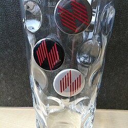 Ein klassisches Dubbeglas aus Glas steht auf einem Tisch und enthält drei runde Buttons mit dem Logo der Hochschule für Wirtschaft und Gesellschaft Ludwigshafen. Die Buttons in Schwarz, Grau und Weiß sind dekorativ an den Einkerbungen des Glases platziert.