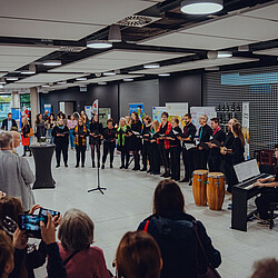 Eine Musikgruppe tritt bei einer Veranstaltung im Foyer auf, begleitet von Klavier und Percussion, während das Publikum zuhört und fotografiert.