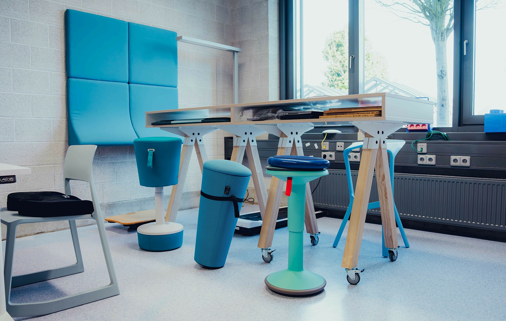 Ein moderner Raum mit flexiblen Sitzmöbeln und höhenverstellbaren Tischen. Das Design kombiniert Holz, Blau- und Grautöne für eine kreative Arbeitsatmosphäre.