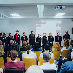 Ein Chor steht vorne im Raum vor einem sitzenden Publikum und präsentiert sich unter dem Titel "Hochschulchor" mit einer Liedprojektion im Hintergrund. Die Teilnehmenden auf der Bühne lächeln und wirken bereit für ihren Auftritt.