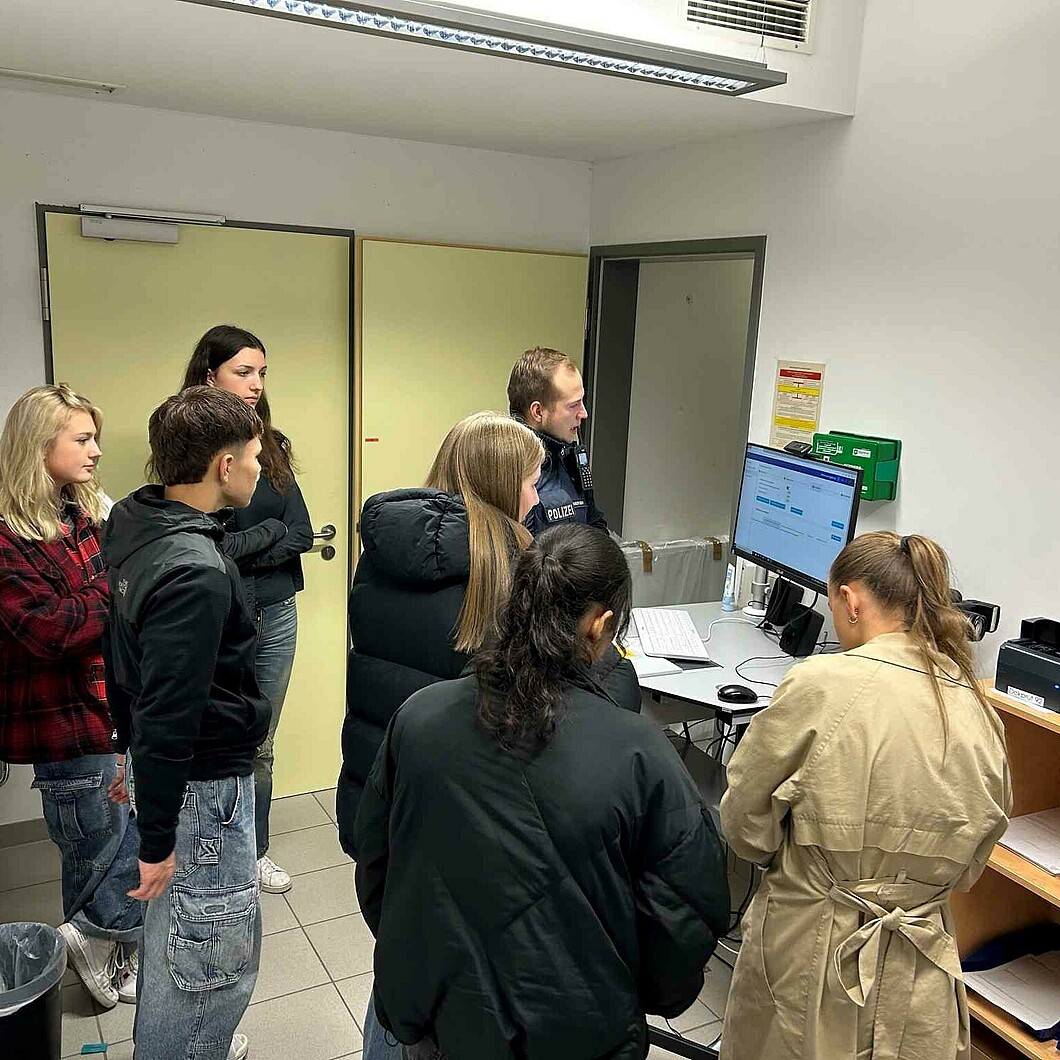 Schüler*innen stehen in einer Polizeistation und verfolgen aufmerksam eine Erklärung eines Polizeibeamten an einem Computerarbeitsplatz.