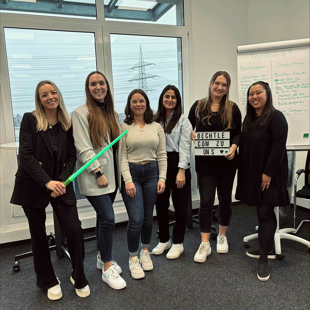 Eine Gruppe junger Frauen posiert in einem modernen Büro mit einem Schild „Bechtle.com zu uns“ – ein humorvolles Detail ist das Lichtschwert in der Hand einer Teilnehmerin.