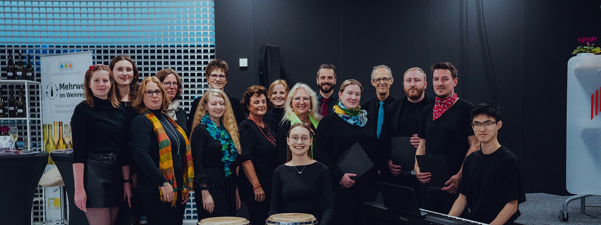 Gruppenbild des Hochschulchors, die Mitglieder tragen einheitlich schwarz  mit einem bunten Akzent. In der Mitte sitzt die Chorleitung Hinter ihr die Mitglieder.
