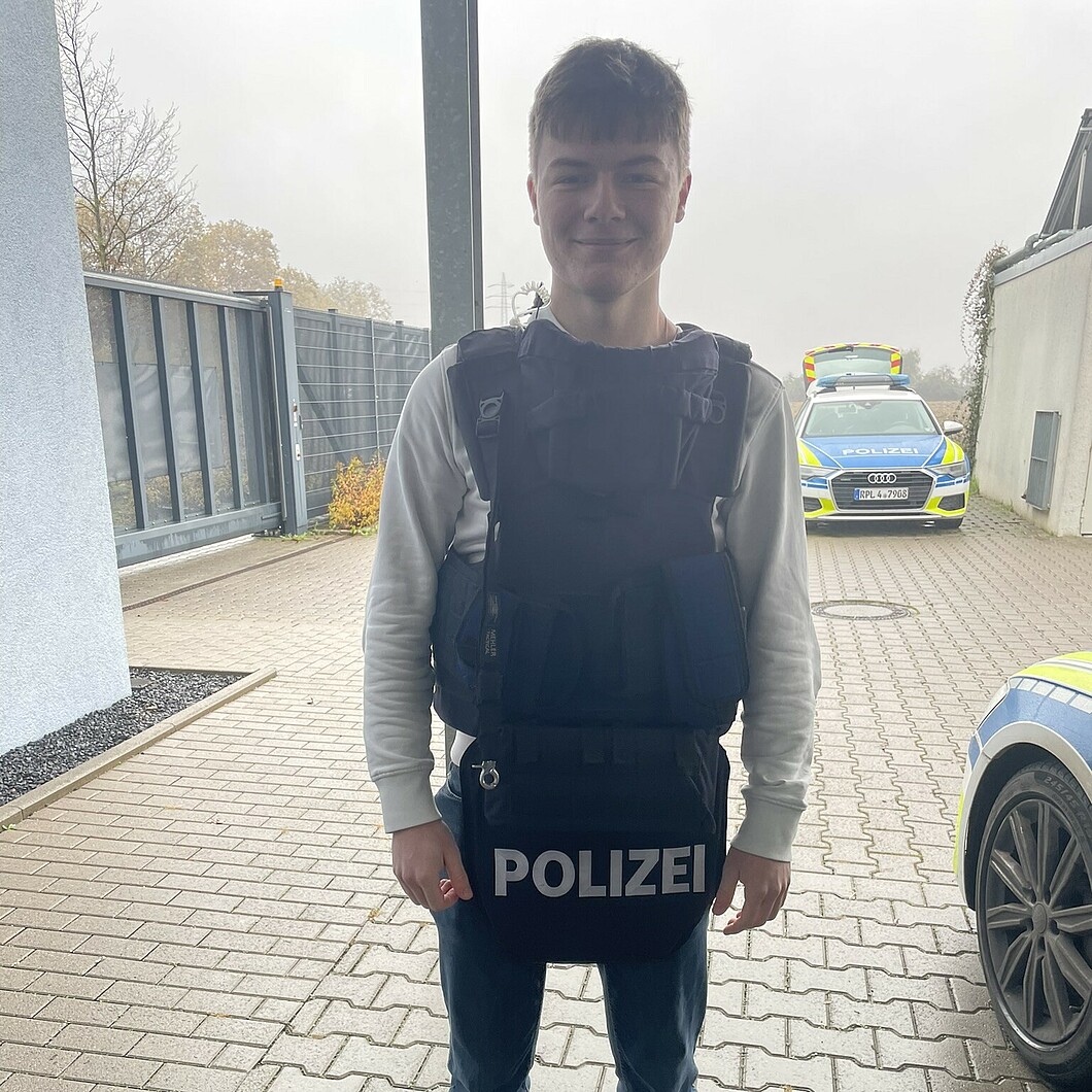 Ein junger Mann trägt eine Schutzweste mit der Aufschrift „Polizei“ und steht lächelnd vor einem Polizeifahrzeug auf einem Außengelände.