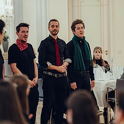  Eine Gruppe von Chorsängern, darunter drei Männer mit bunten Krawatten und Schals, singt zusammen vor dem Publikum.