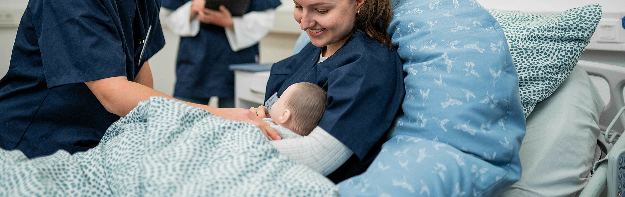Drei Studentinnen der Hebammenwissenschaft stellen mit einer Säuglingspuppe eine Szene auf der Wöchnerinnenstation nach