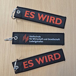 Drei schwarze Textil-Schlüsselanhänger mit Metallring liegen auf einem Tisch, zwei zeigen in roter Schrift den Aufdruck „ES WIRD“, einer trägt zusätzlich das Hochschullogo. Die Anhänger wirken stabil und sind hochwertig verarbeitet.