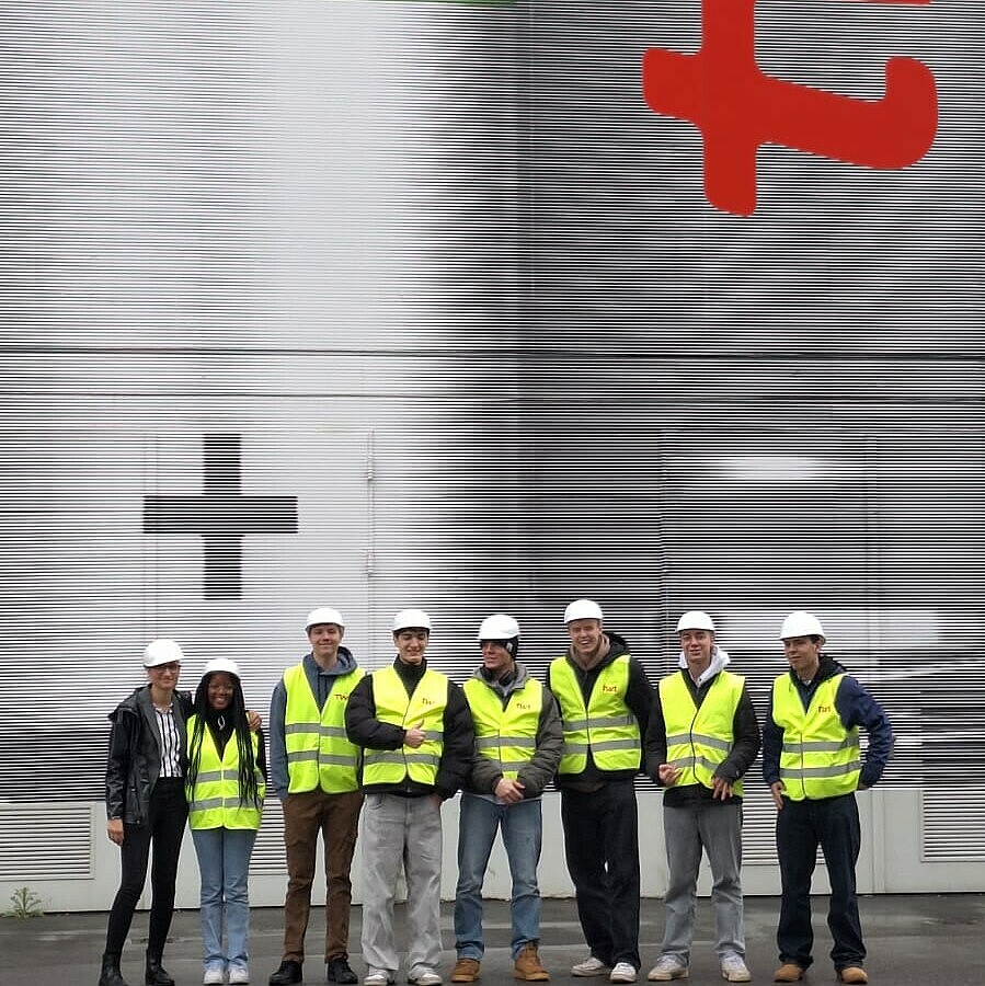 Eine Gruppe junger Menschen trägt Schutzwesten und Helme vor einer großen Industrieanlage mit dem Logo „TWA“, was auf einen Besuch bei einem Produktionsbetrieb hinweist.